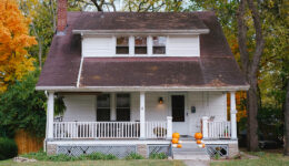 Small white house set in the trees with pumpkins on the front steps.